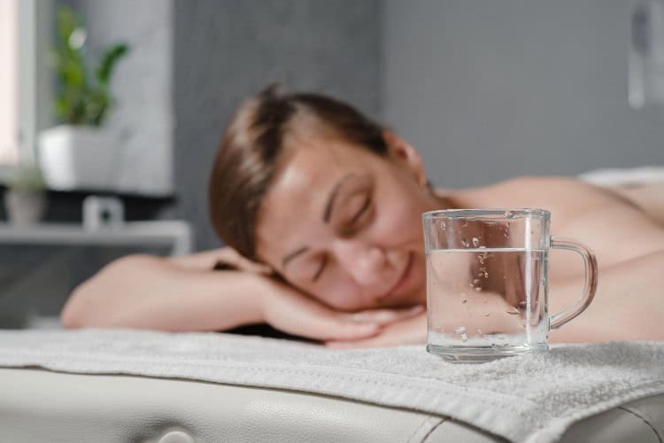 Relaxed woman preparing for massage with water beside bed